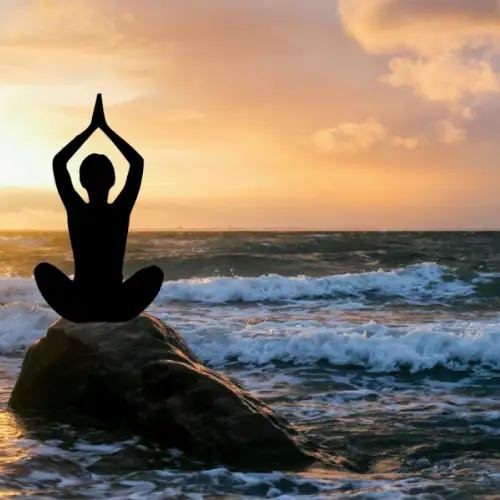 A silhouetted person meditating in a lotus position on a seaside rock at sunset, with ocean waves surrounding them and a golden sky in the background.