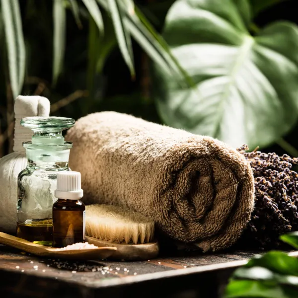 A relaxing spa setup featuring a rolled towel, essential oils, dried lavender, a brush, and salt, surrounded by lush greenery.