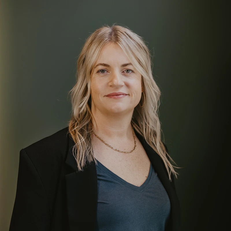 A professional headshot of Angie Gignac, owner of Elements Skin Care Spa. She has blonde, wavy hair, wearing a black blazer and blue top, smiling against a dark green background.