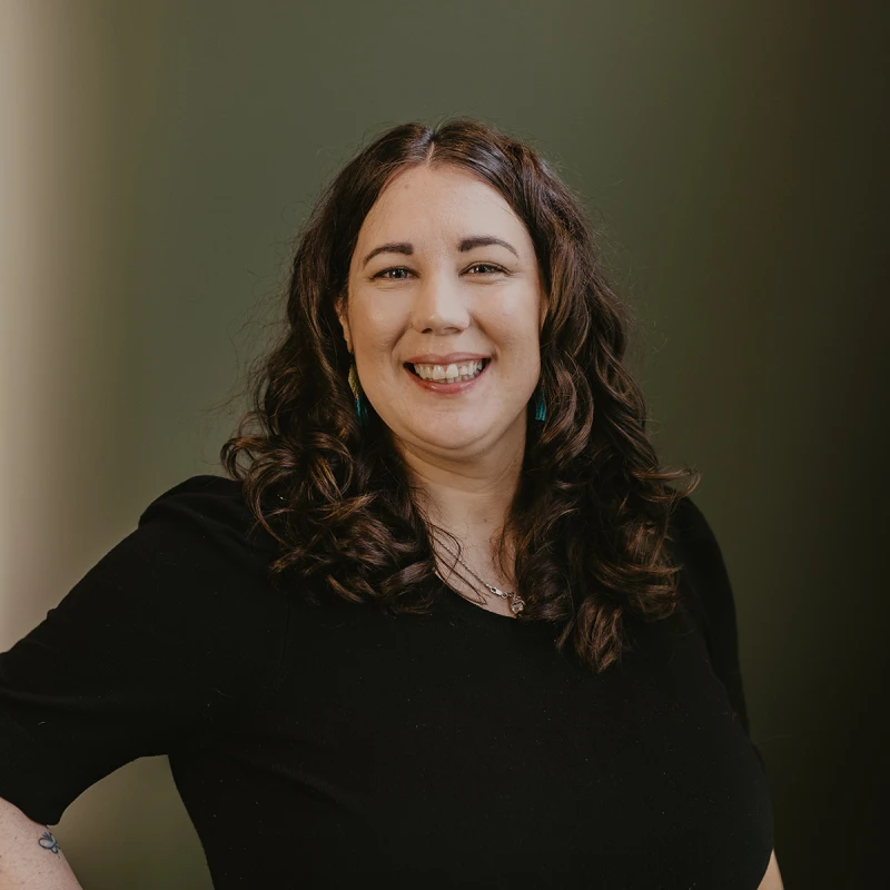 A portrait of Carrissa Busch wearing a black top and turquoise earrings, smiling warmly against a soft green background. Her dark hair is styled in loose curls, and she exudes a friendly, approachable demeanour.