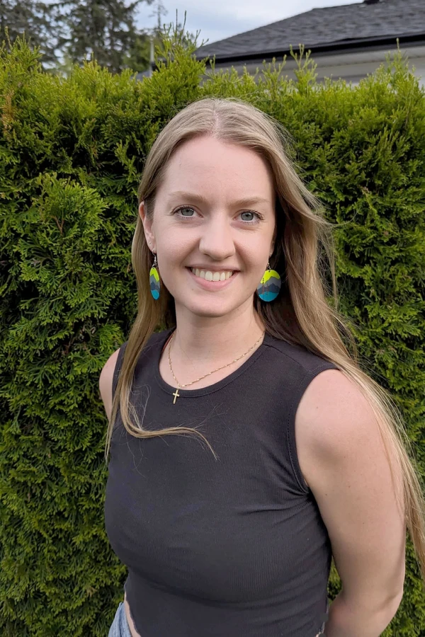 Marissa Berger, massage wellness specialist at Elements Spa in Nanaimo, BC. She is smiling and standing outdoors in front of an evergreen hedge. She is wearing colourful circular earrings and a black tank top.