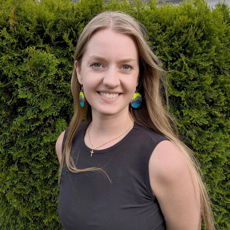 Marissa Berger, massage wellness specialist at Elements Spa in Nanaimo, BC. She is smiling and standing outdoors in front of an evergreen hedge. She is wearing colourful circular earrings and a black tank top.