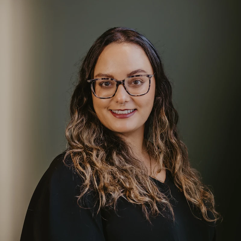A professional portrait of a Skylar Byman, Aesthetician. She has shoulder-length curly hair and is wearing glasses and a black outfit. The background is dark green and subtly illuminated.