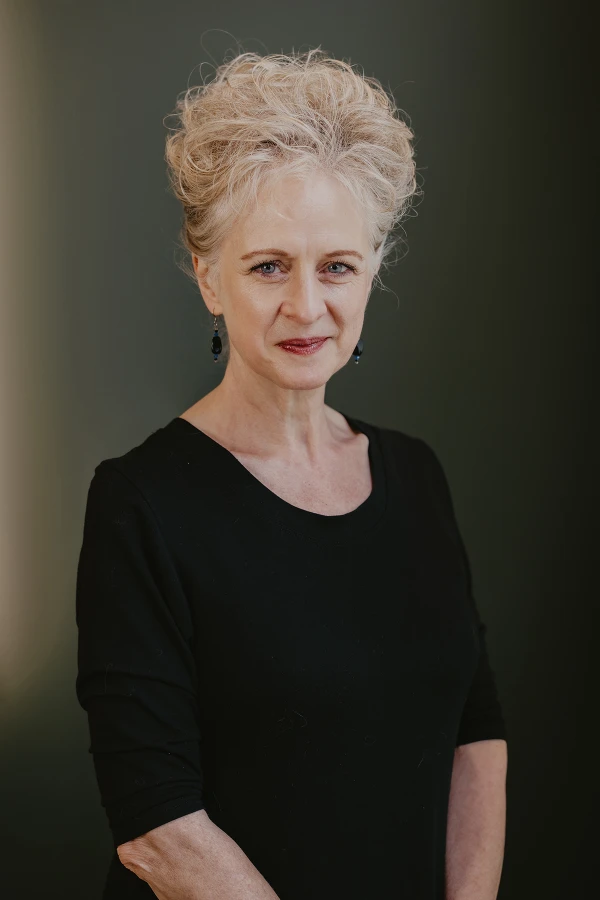 A portrait of Tina Roselle. She has short, curly blonde hair, and is wearing a black top and subtle makeup, looking intently at the camera against a plain green background.
